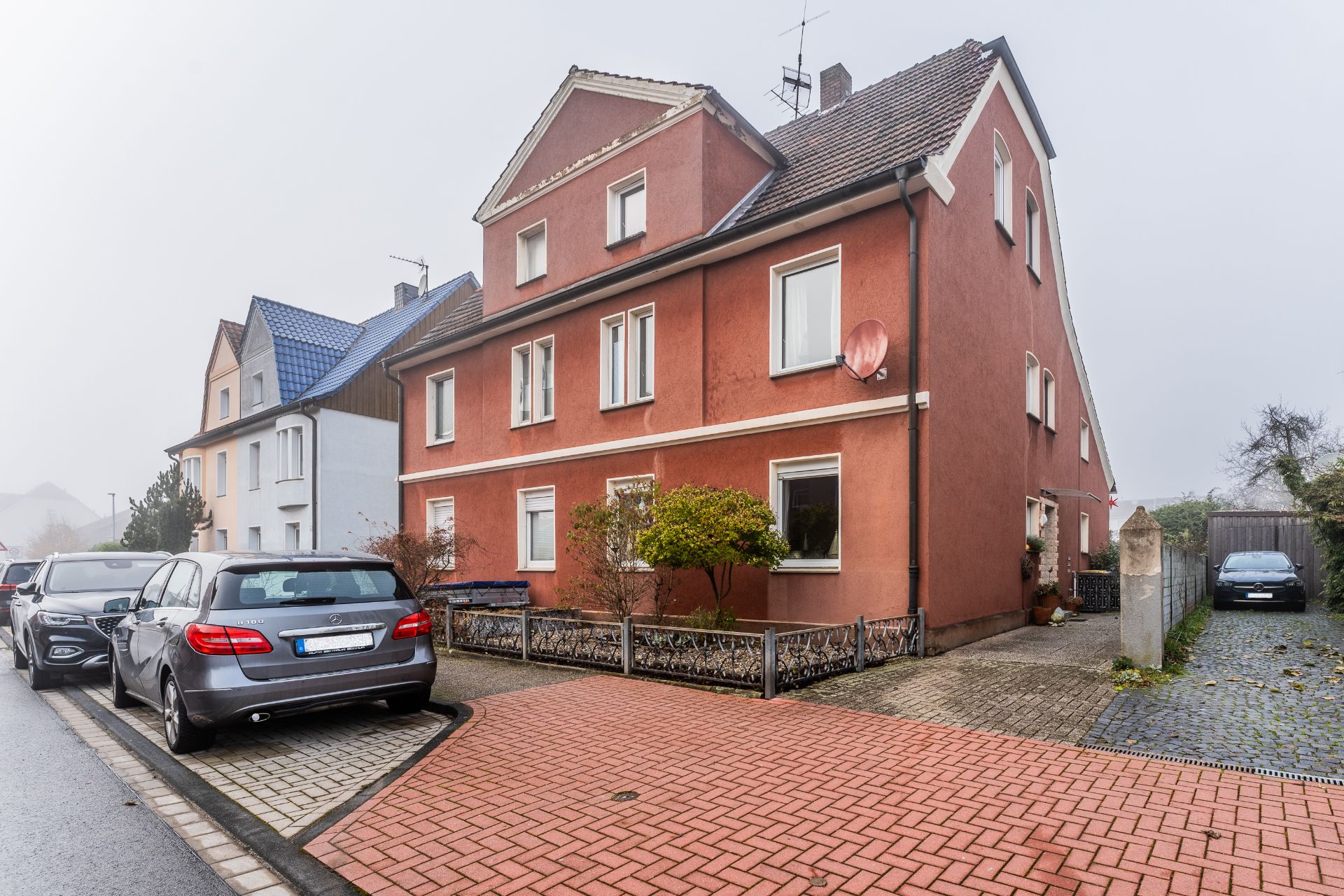Rotes Wohnhaus mit mehreren Fenstern, geparkten Autos und kleinem Vorgarten.
