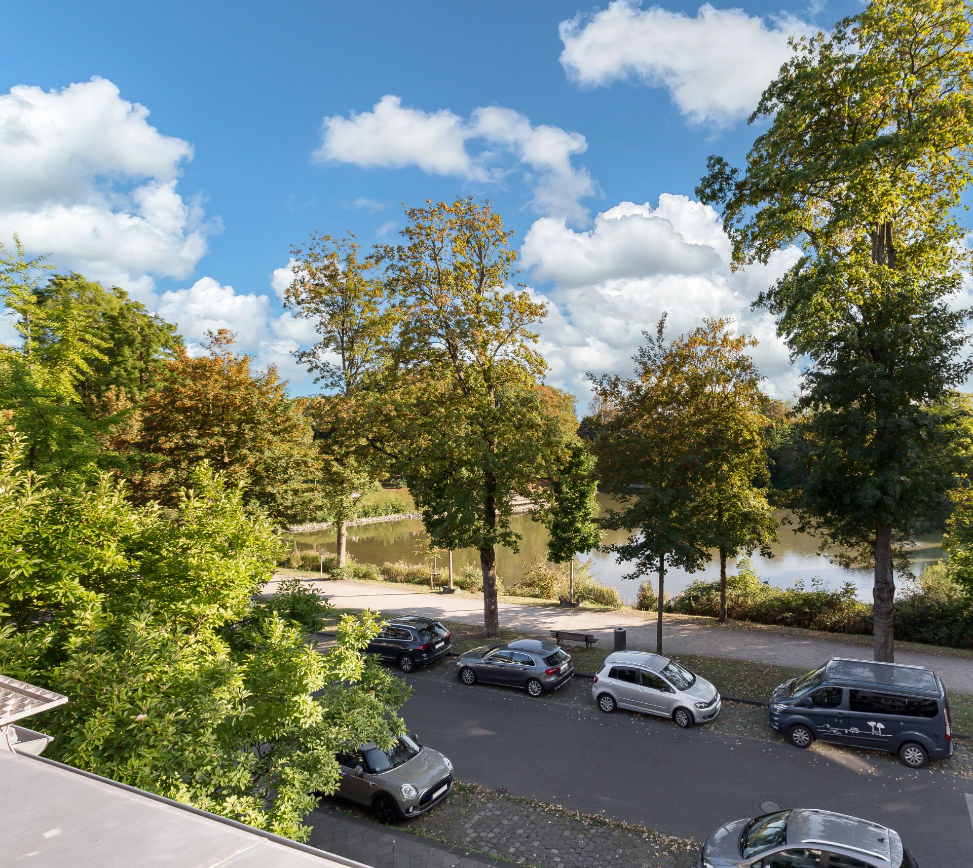 Blick auf eine Baumallee mit geparkten Autos, dahinter ein See und blauer Himmel mit Wolken.