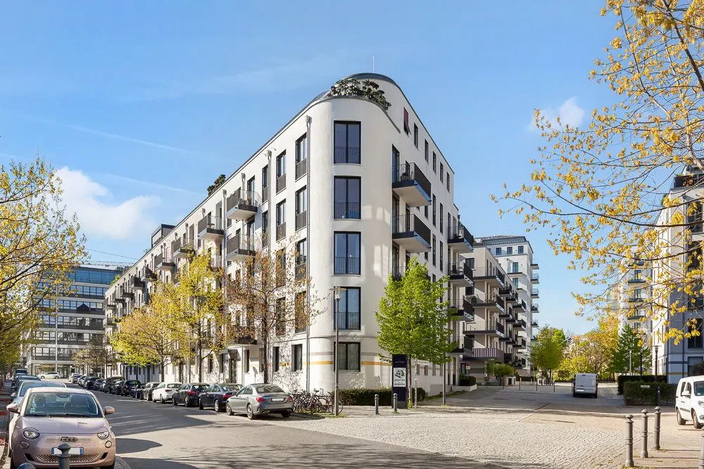 Modernes Wohngebäude mit Balkonen an einer Straßenecke, Bäume und parkende Autos davor.