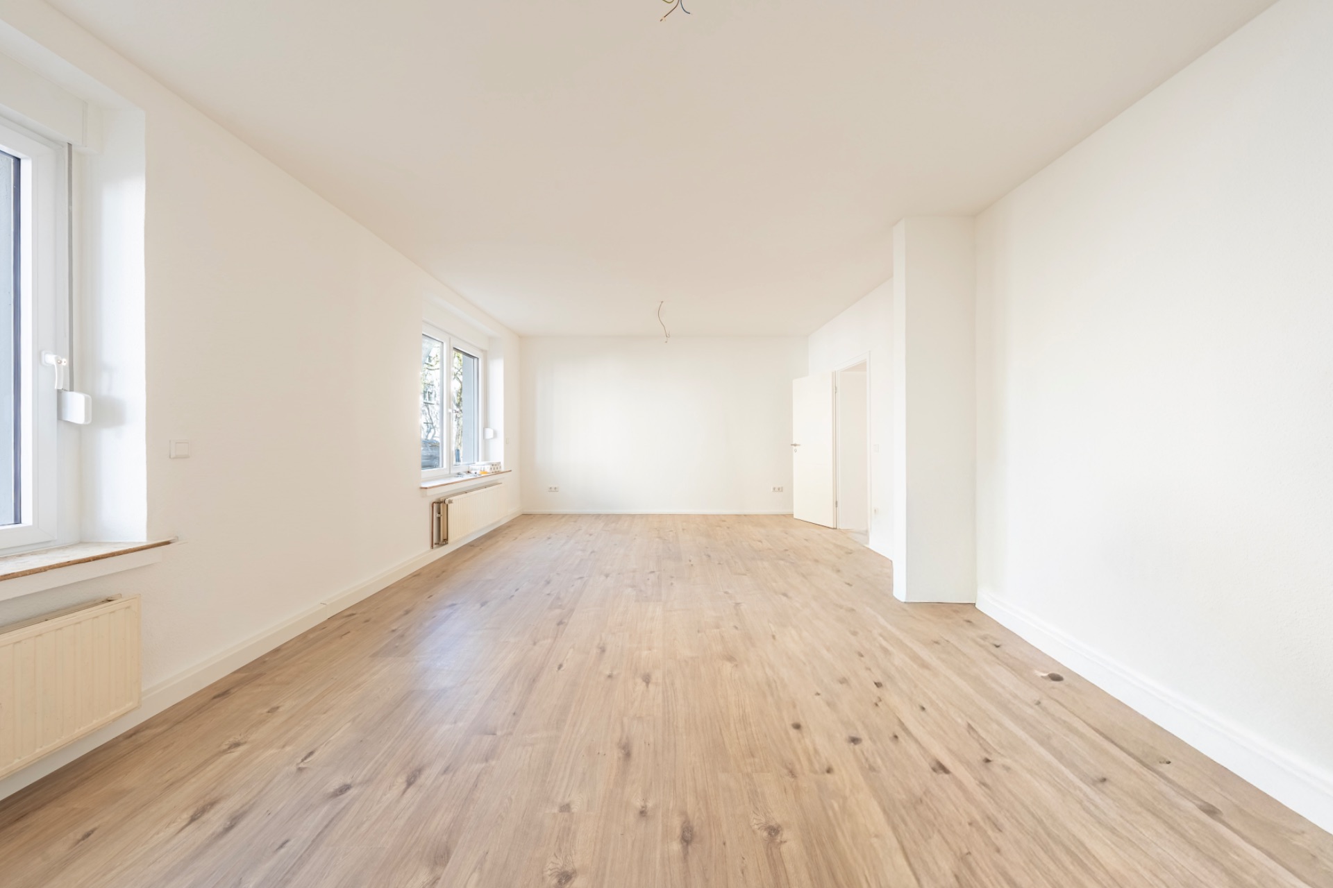 Helles, leeres Zimmer mit Holzfußboden, weißen Wänden und zwei Fenstern.