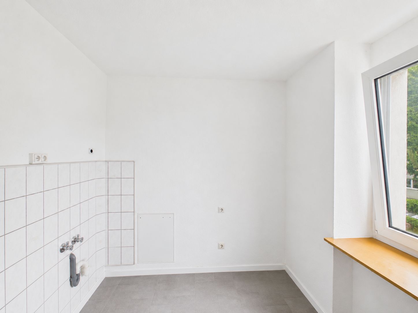 Leerer, weißer Raum mit Fliesenspiegel, Wasseranschlüssen und offenem Fenster.