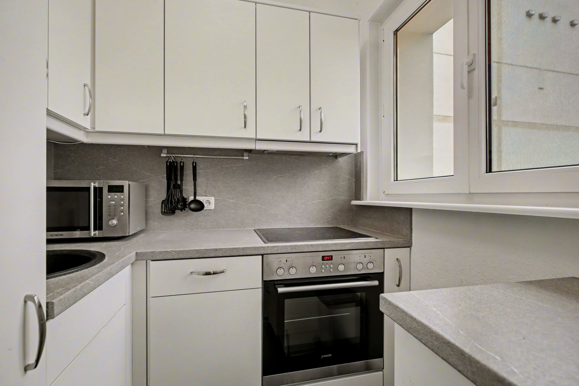 Moderne, kleine Küche mit weißen Schränken, Herd, Backofen, Mikrowelle und großem Fenster.