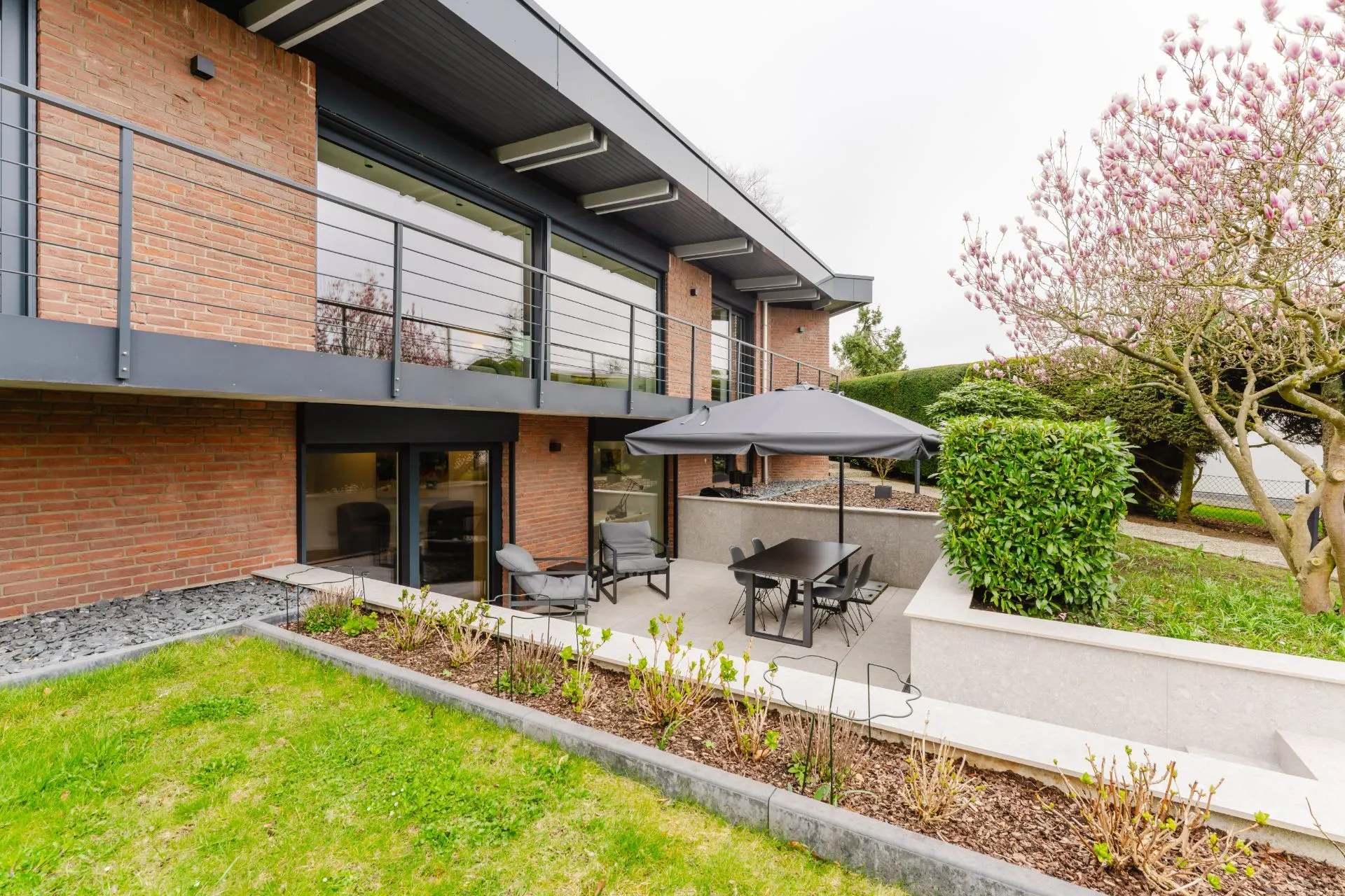 Moderne Terrasse mit Gartenmöbeln, Sonnenschirm und blühendem Baum an einem Wohnhaus.
