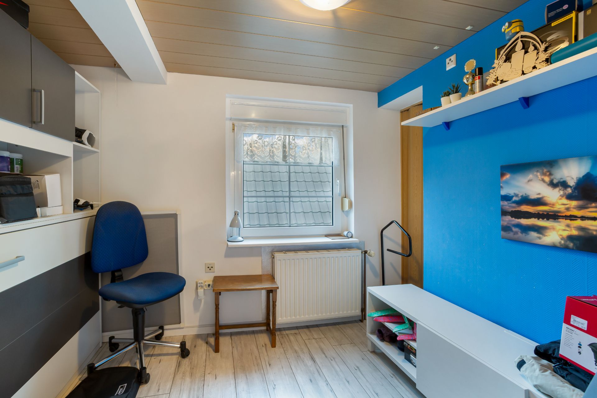 Kleines Arbeitszimmer mit Schreibtischstuhl, Regal, Fenster, Heizkörper und blauer Wand mit Bild.