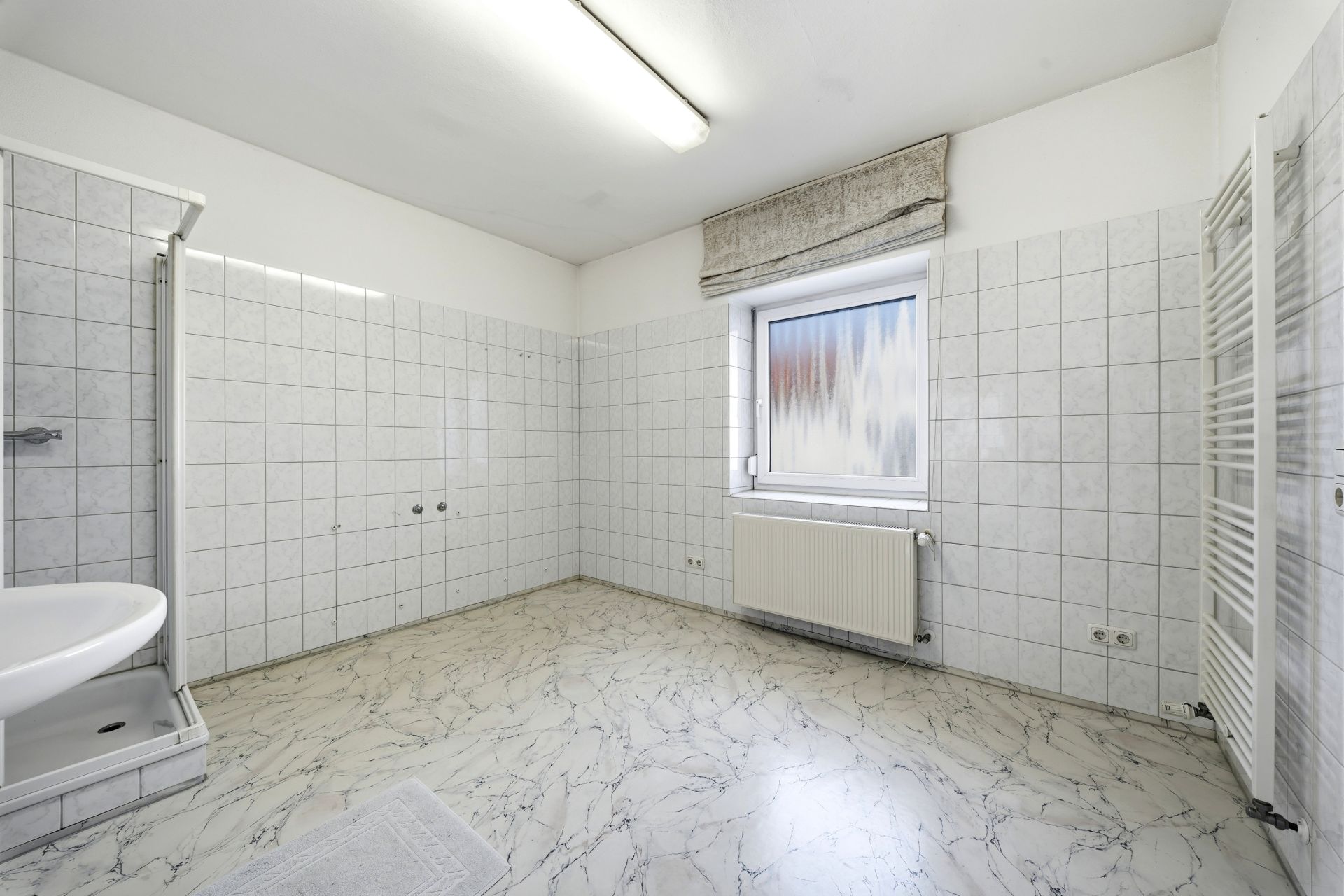 Leeres, helles Badezimmer mit Fliesen, Fenster, Waschbecken, Dusche und Handtuchheizkörper.
