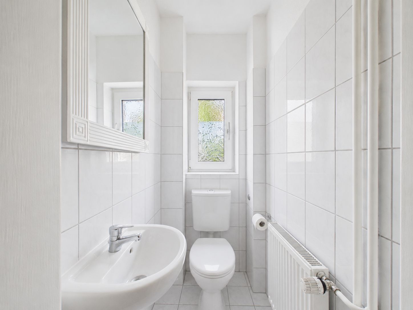 Helles, schmales Badezimmer mit Waschbecken, Toilette, Spiegel und Fenster.
