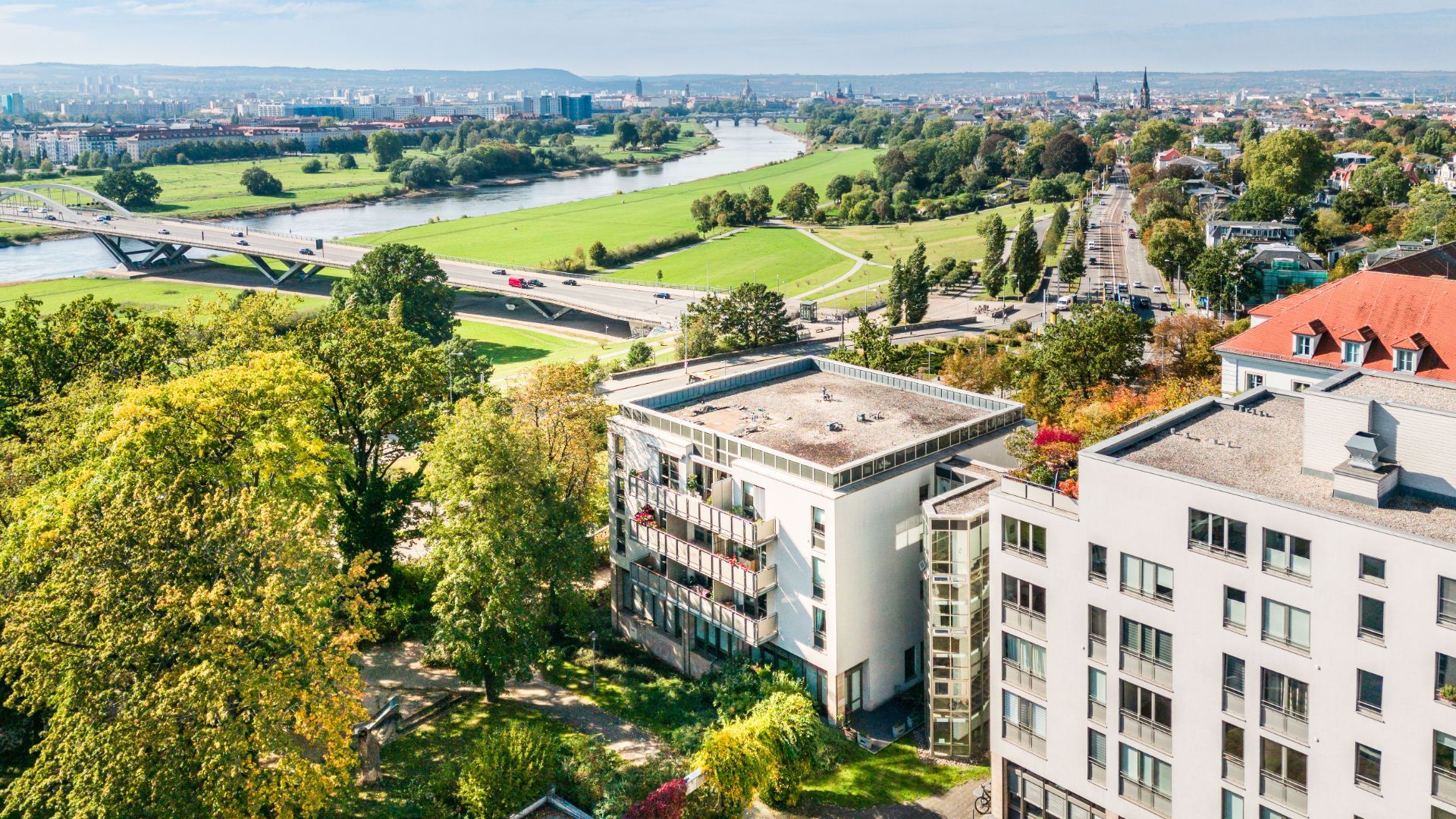 Wohnhäuser mit Balkonen, Bäumen und Blick auf einen Fluss mit Brücke im Hintergrund.