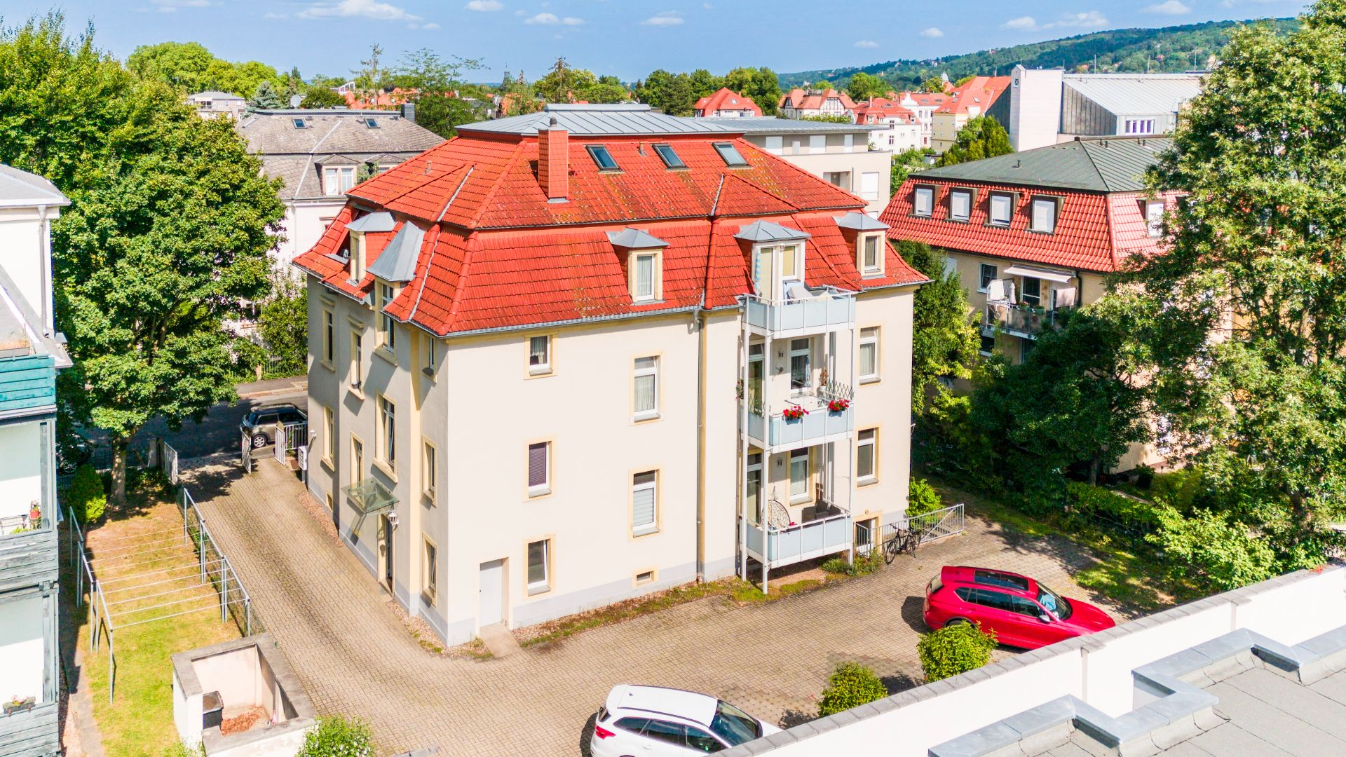 Mehrfamilienhaus mit rotem Dach, Balkonen und zwei parkenden Autos vor dem Gebäude.
