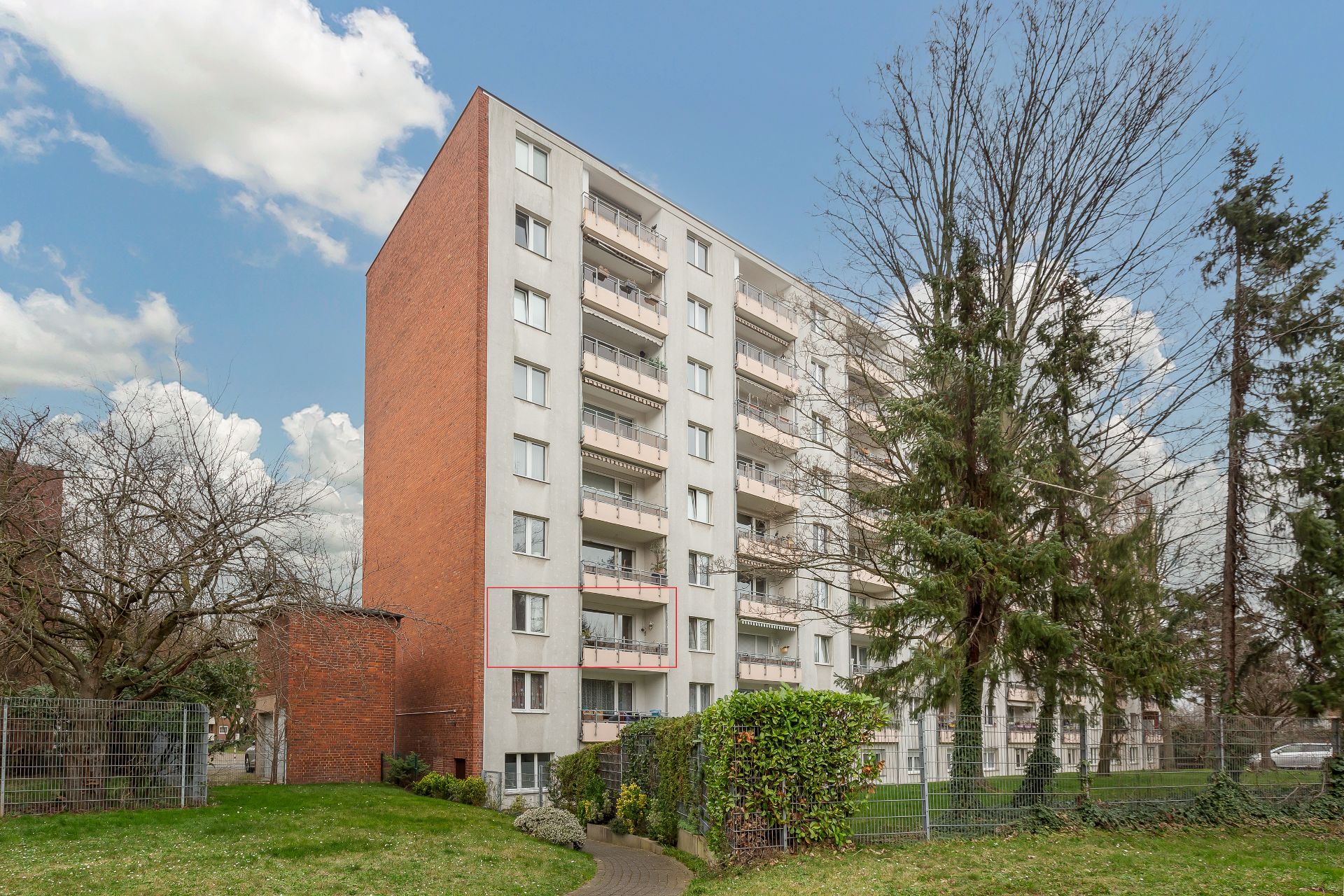 Mehrstöckiges Wohnhaus mit Balkonen und grünem Vorgarten, daneben Bäume und ein Zaun.