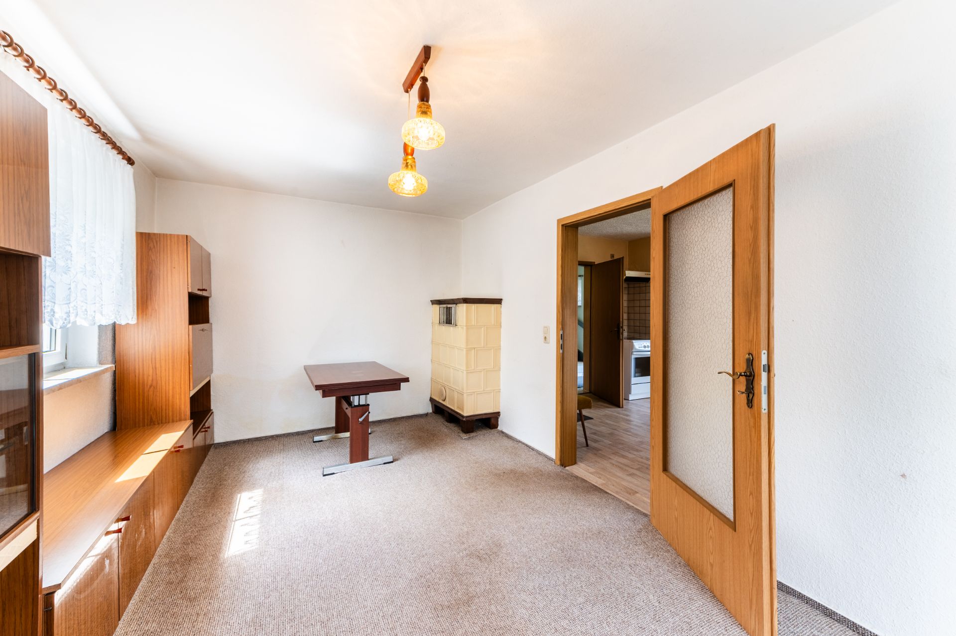 Helles Wohnzimmer mit Teppichboden, Holzschrank, kleinem Tisch und offener Tür zum nächsten Raum.