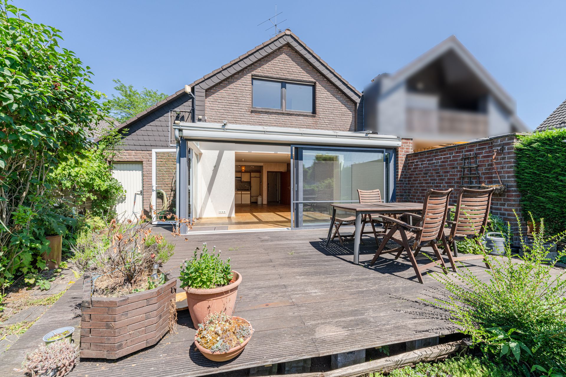 Holzterrasse mit Tisch und Stühlen vor einem Einfamilienhaus, Pflanzen im Vordergrund.