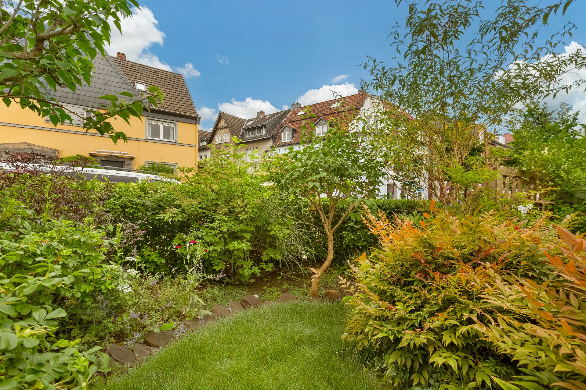 Gepflegter Garten mit verschiedenen Sträuchern und Bäumen, im Hintergrund Wohnhäuser.