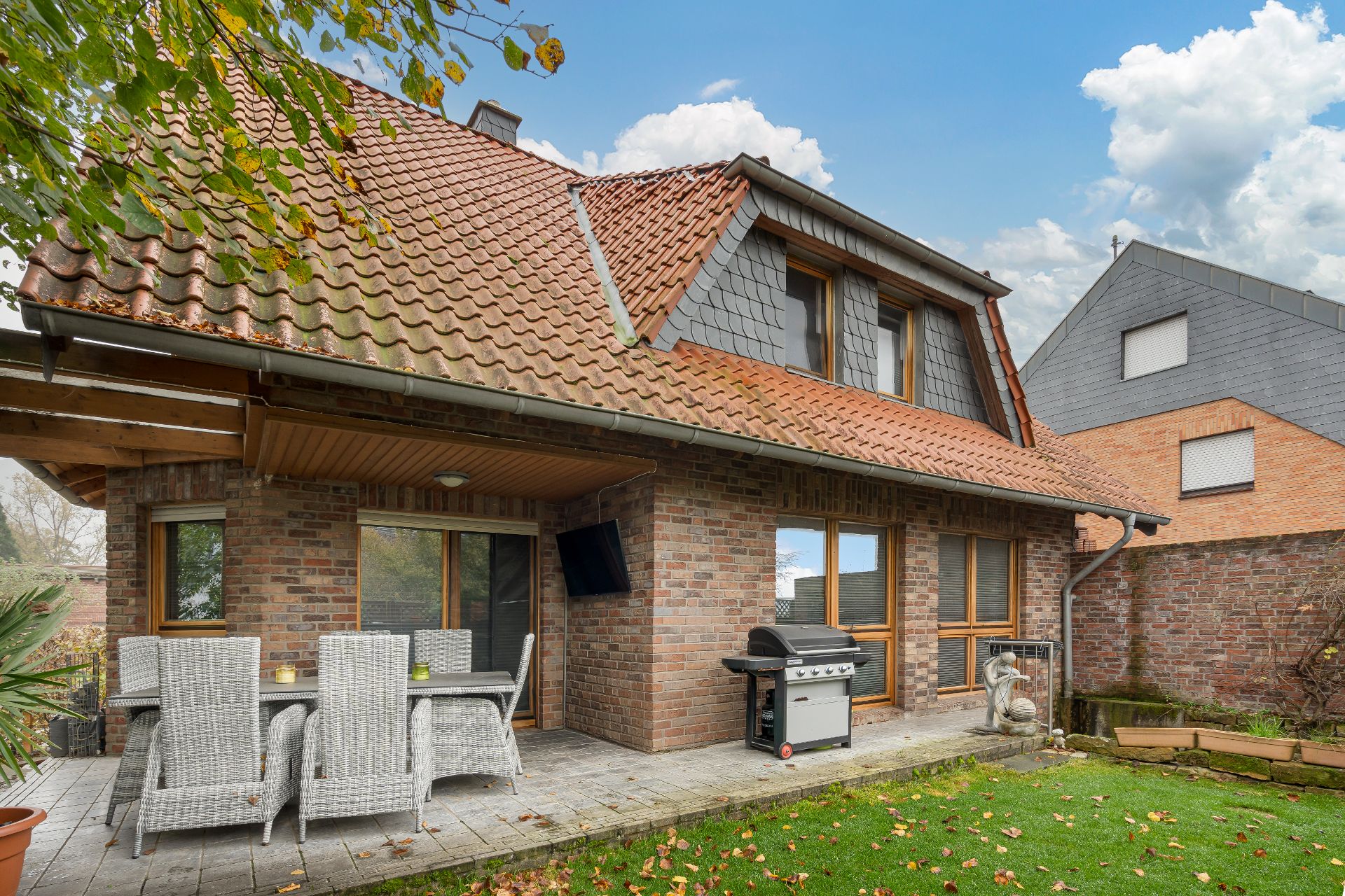 Backsteinhaus mit Terrasse, Gartenmöbeln, Grill und kleinem Rasen im Vordergrund.