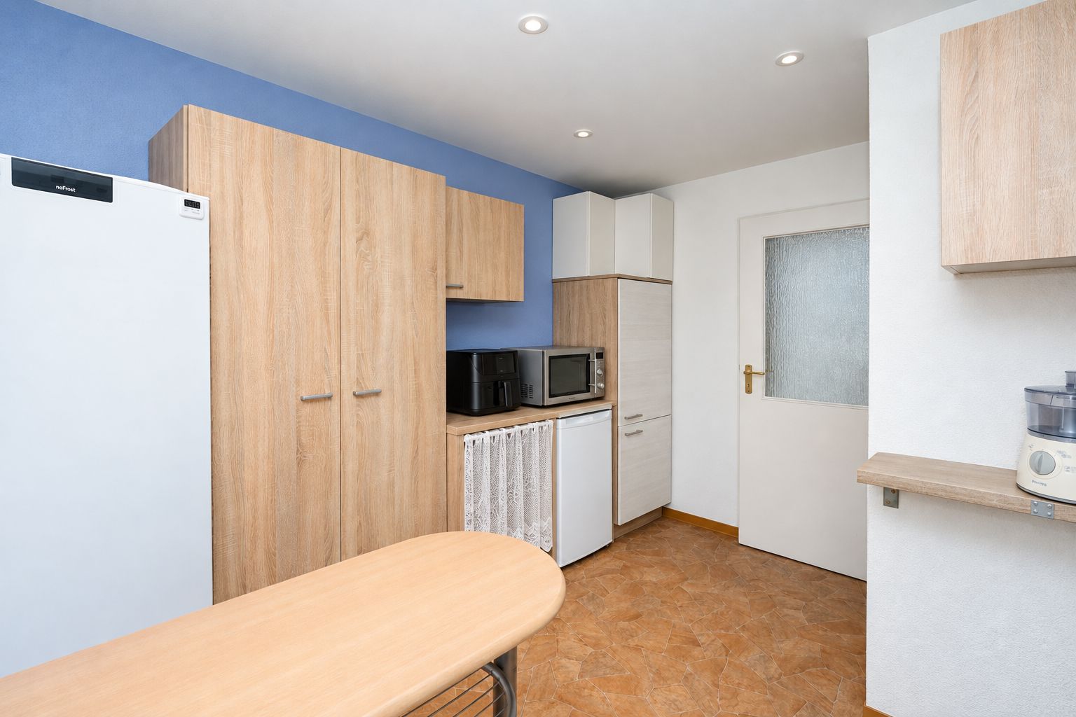Moderne Küche mit Holzmöbeln, Kühlschrank, Mikrowelle und heller Tür.