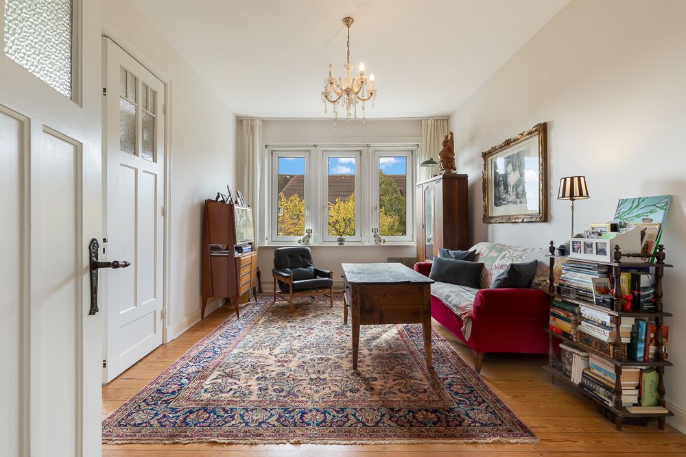 Helles Wohnzimmer mit Teppich, rotem Sofa, Schreibtisch, Regal und großen Fenstern.