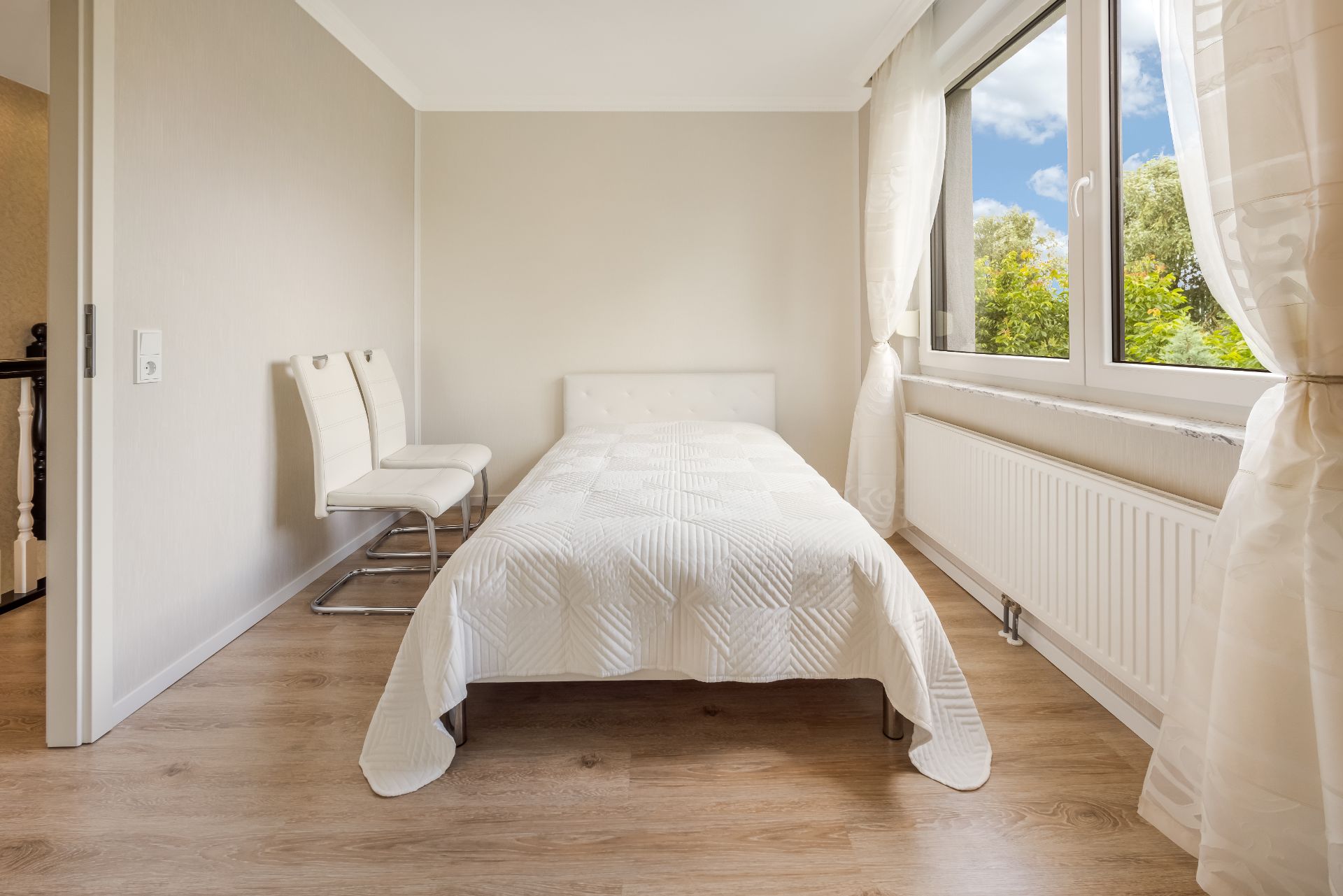 Helles Schlafzimmer mit Einzelbett, zwei weißen Stühlen und großem Fenster mit Blick ins Grüne.