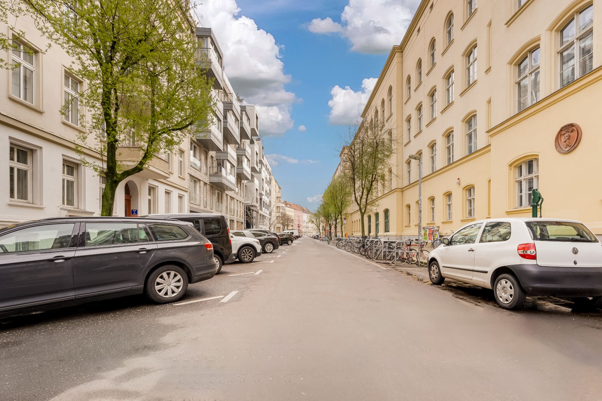 Ruhige Wohnstraße mit geparkten Autos, Fahrrädern und Bäumen an beiden Seiten.