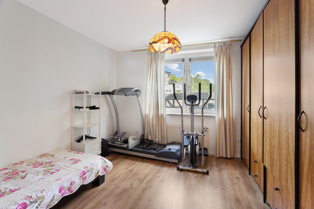 Zimmer mit Bett, Fitnessgeräten, Regal, Kleiderschrank und Fenster mit Vorhängen.