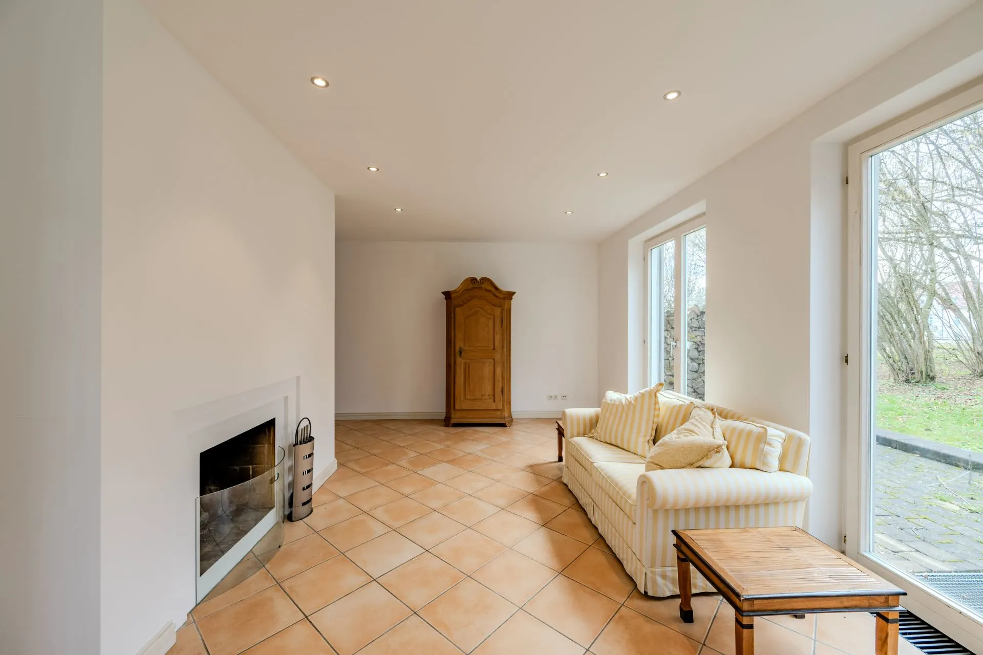 Helles Wohnzimmer mit Kamin, Sofa, Holztisch und Schrank, große Fenster mit Blick nach draußen.