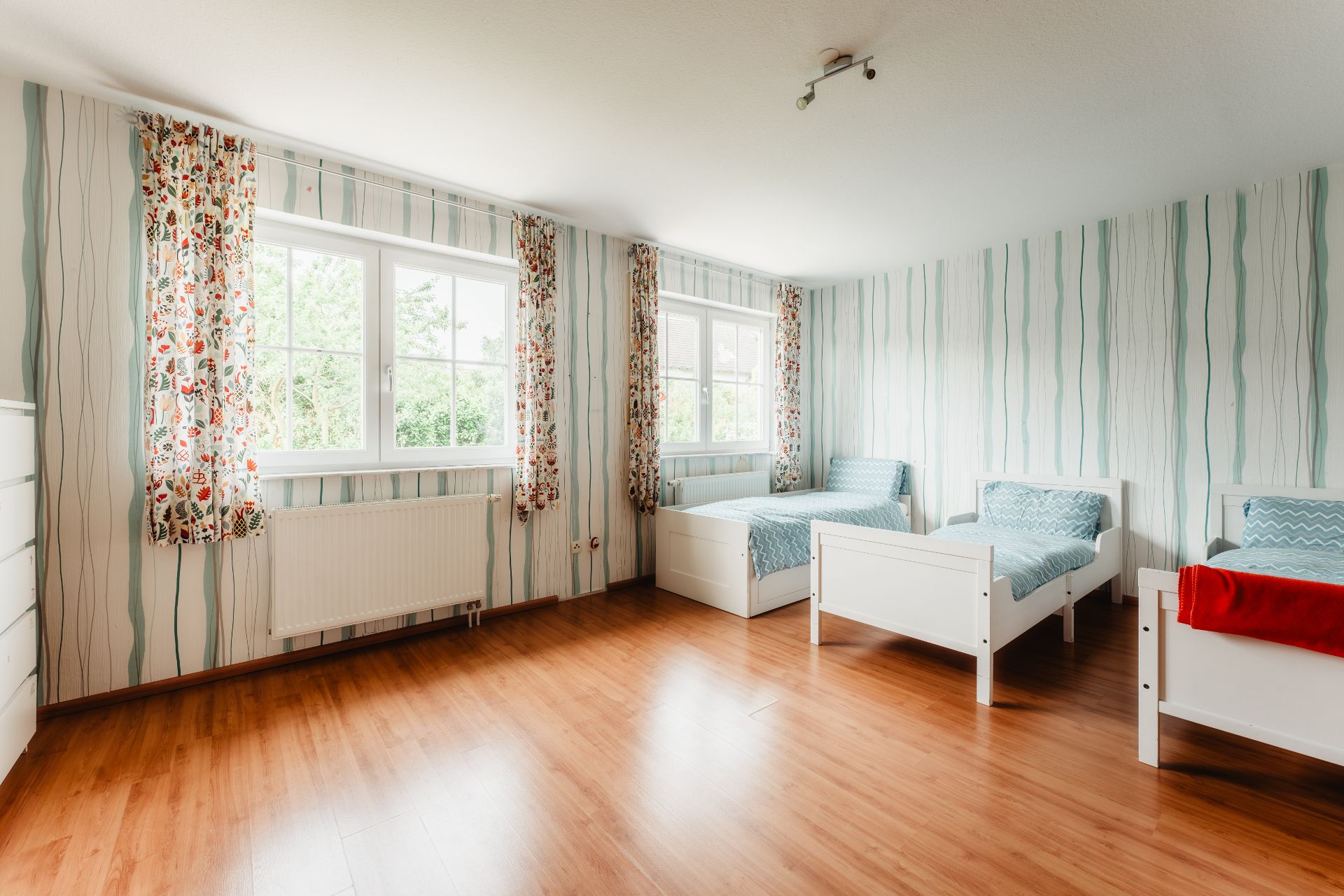 Helles Schlafzimmer mit drei Einzelbetten, Holzfußboden und zwei großen Fenstern mit bunten Vorhängen.
