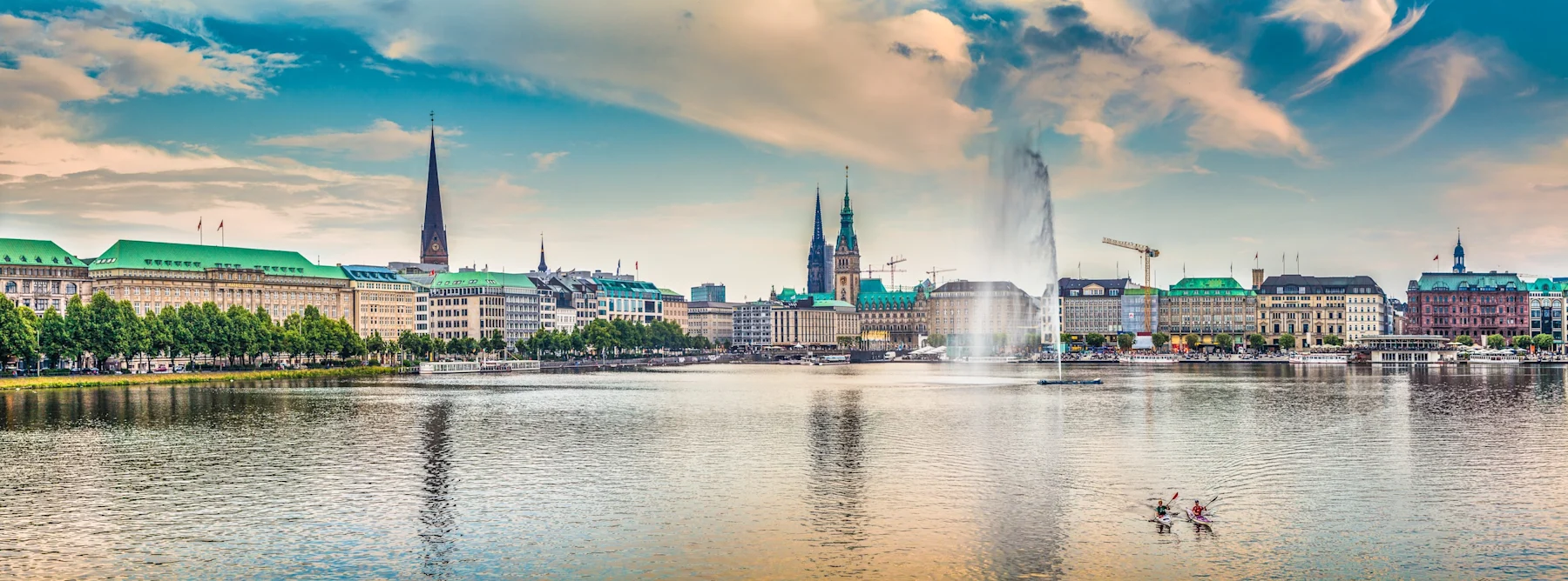Blick über einen großen See mit Springbrunnen, umliegenden Gebäuden und Kirchen im Hintergrund.