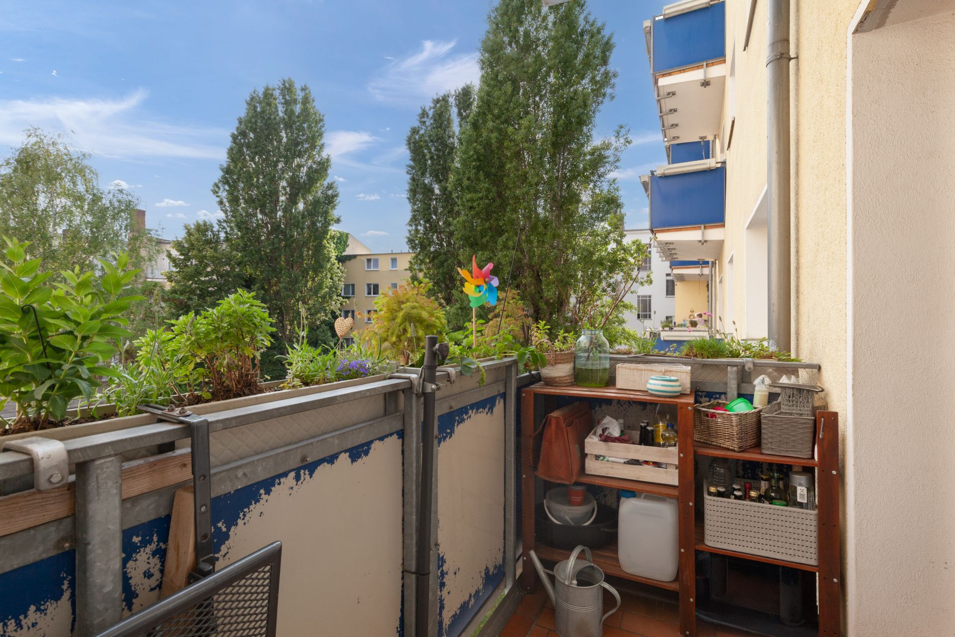 Kleiner Balkon mit Pflanzen, Regal und Blick auf Bäume sowie benachbarte Wohngebäude.
