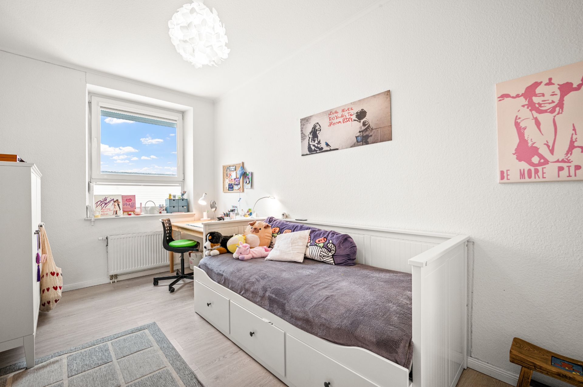 Helles Kinderzimmer mit Einzelbett, Schreibtisch am Fenster und Kuscheltieren auf dem Bett.