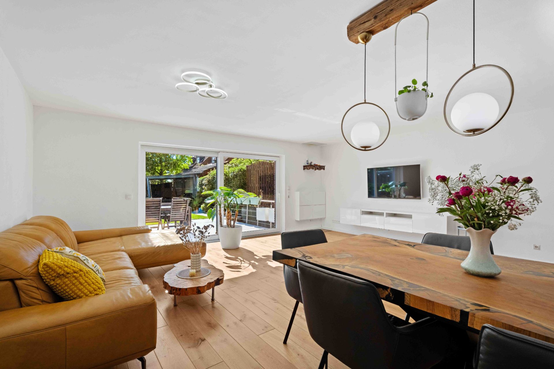 Helles Wohnzimmer mit Leder-Ecksofa, Holztisch, TV, Blumen und großem Fenster zum Garten.