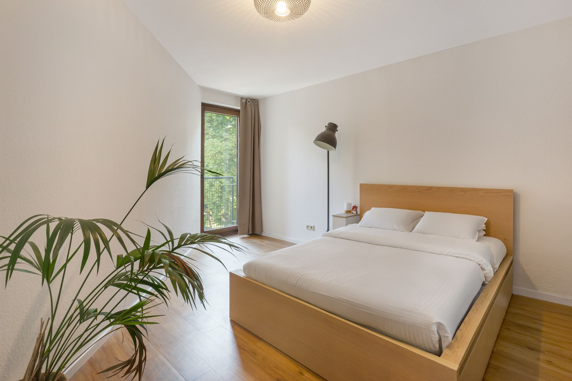 Helles Schlafzimmer mit großem Bett, Holzfußboden, Stehlampe und einer Pflanze am Fenster.