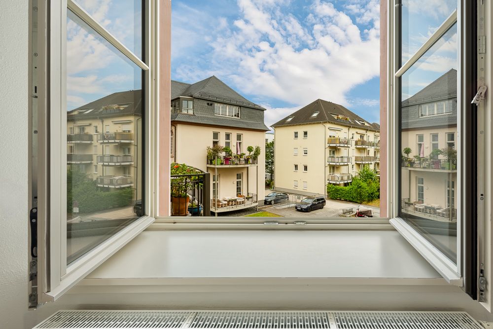 Blick durch ein offenes Fenster auf mehrere Wohngebäude mit Balkonen und geparkten Autos.