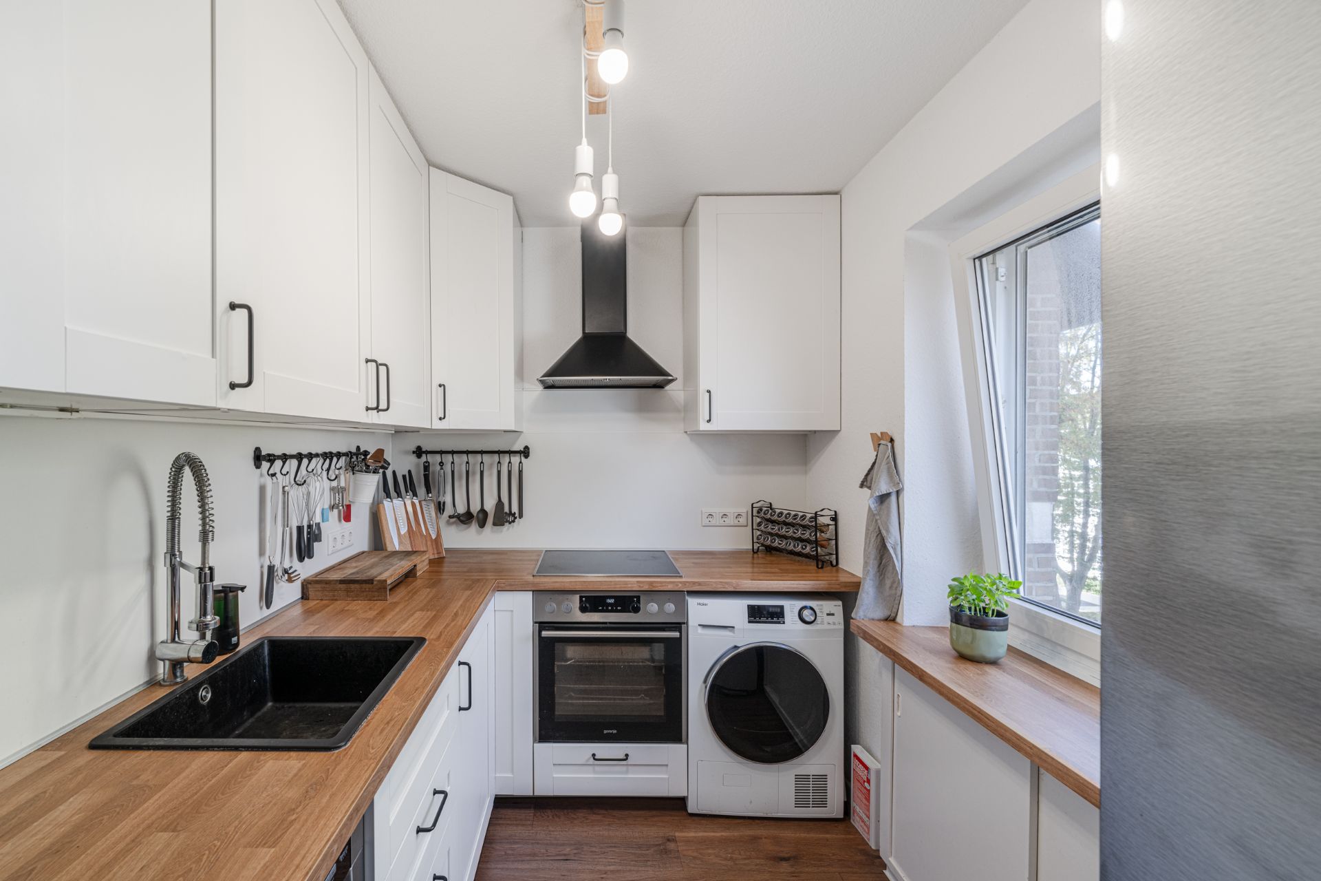 Moderne Küche mit weißen Schränken, Holzarbeitsplatte, Herd, Spüle, Waschmaschine und großem Fenster.