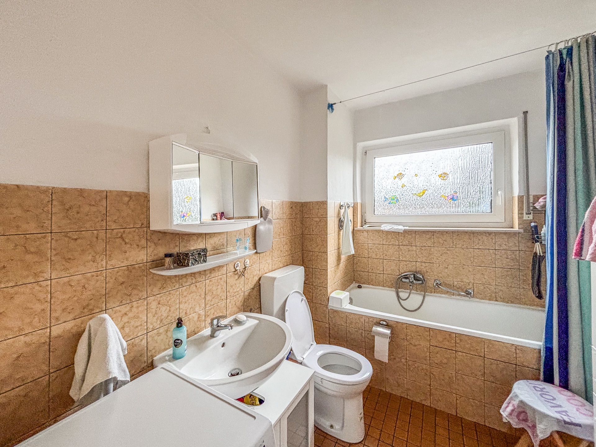 Helles Badezimmer mit Badewanne, Waschbecken, Toilette, Spiegel und Fenster mit bunten Stickern.