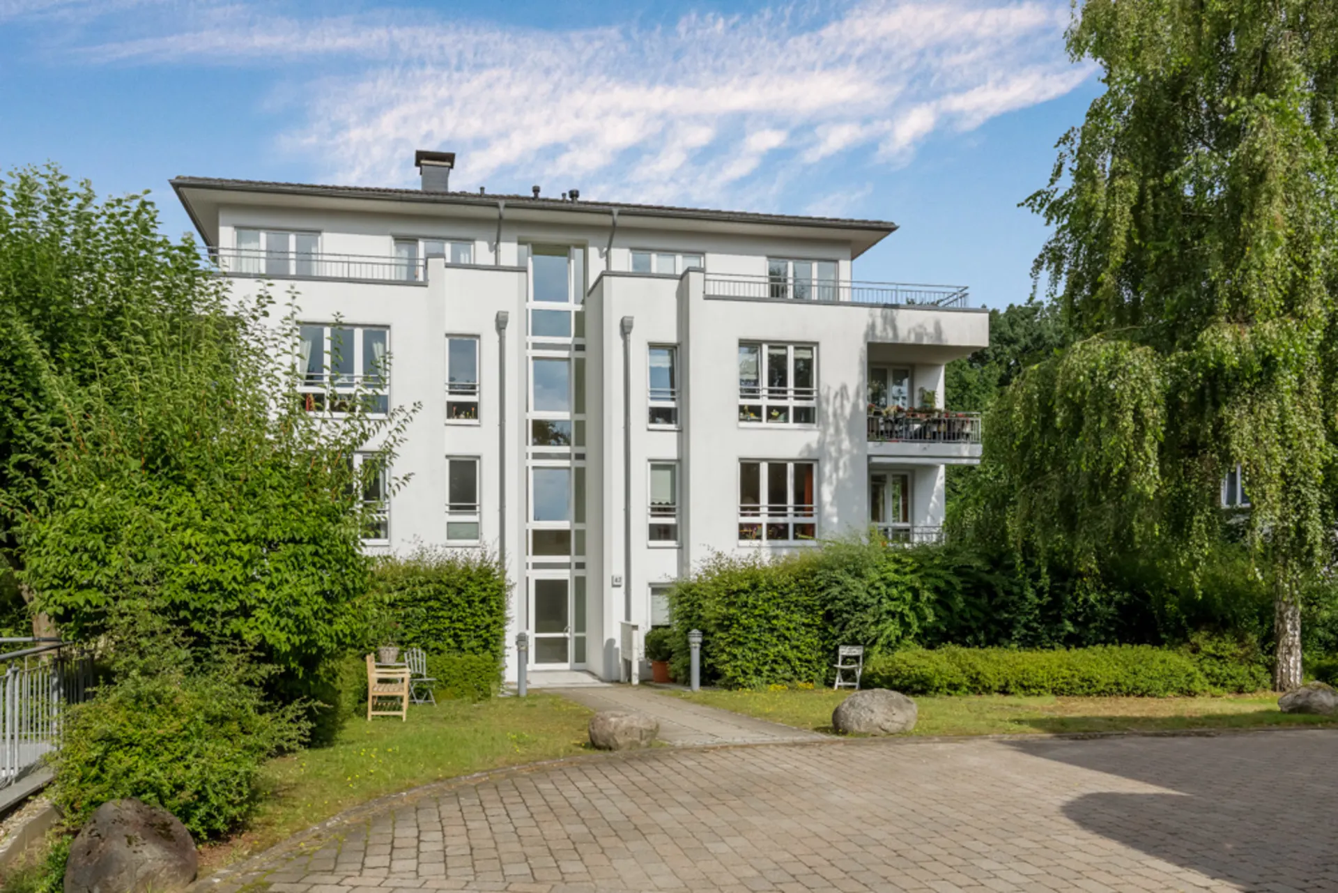 Modernes, weißes Wohnhaus mit drei Etagen, großen Fenstern und umgeben von viel Grün.