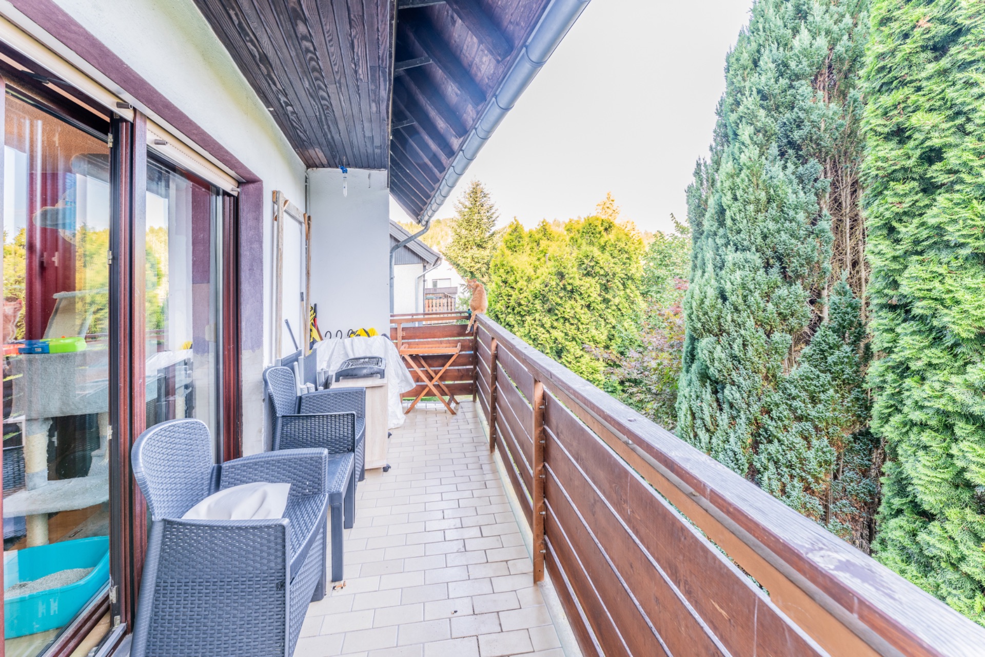 Langer Balkon mit Sitzgelegenheiten, Holzzaun und Blick auf dichte grüne Bäume.