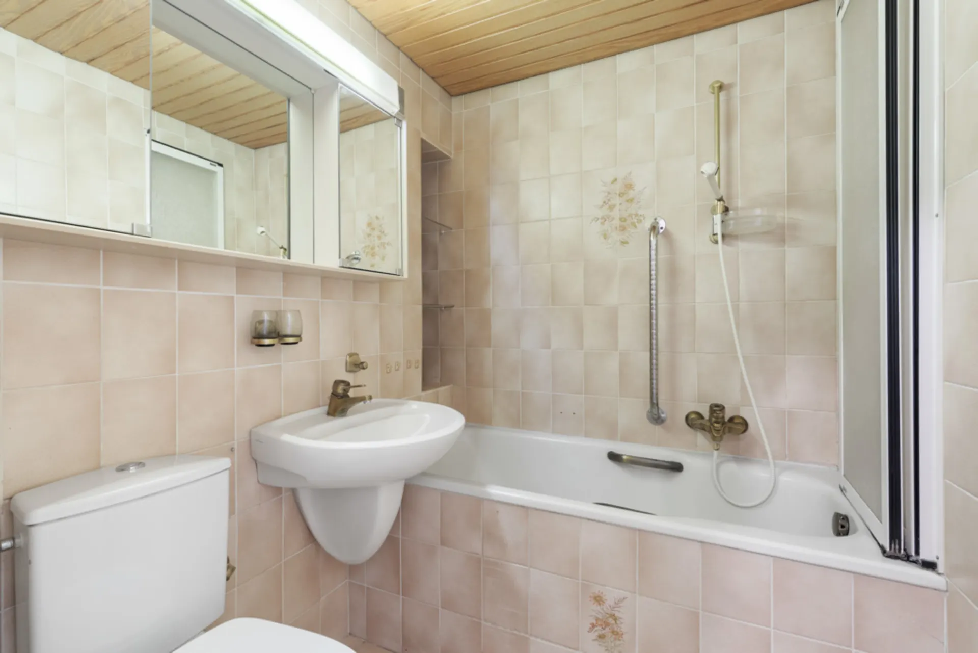 Kleines Badezimmer mit Badewanne, Waschbecken, Spiegel und WC, beige Fliesen und Holzdecke.