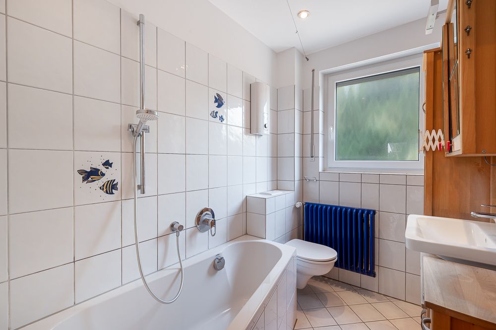 Helles Badezimmer mit Badewanne, WC, Waschbecken und Fenster, weiße Fliesen mit Fischmotiven.