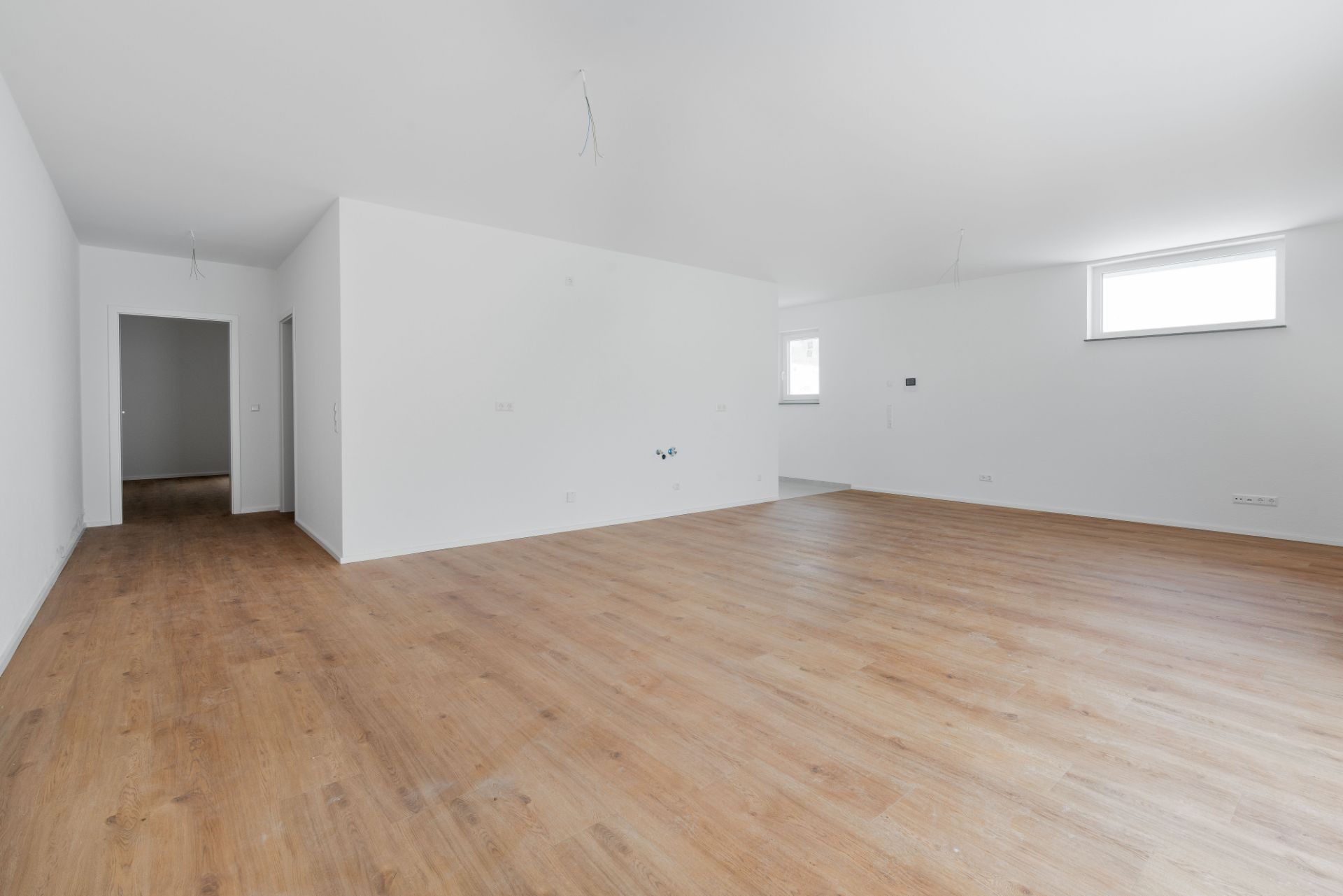 Heller, leerer Raum mit Holzfußboden und weißen Wänden, zwei Fenster und eine offene Tür sichtbar.