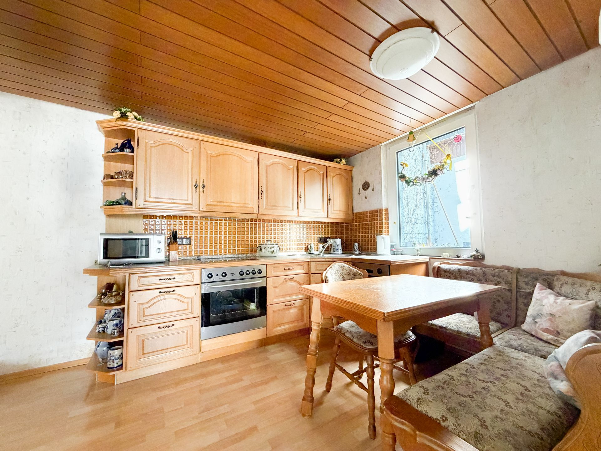 Holzküche mit Eckbank, Tisch, Backofen, Mikrowelle und großem Fenster.