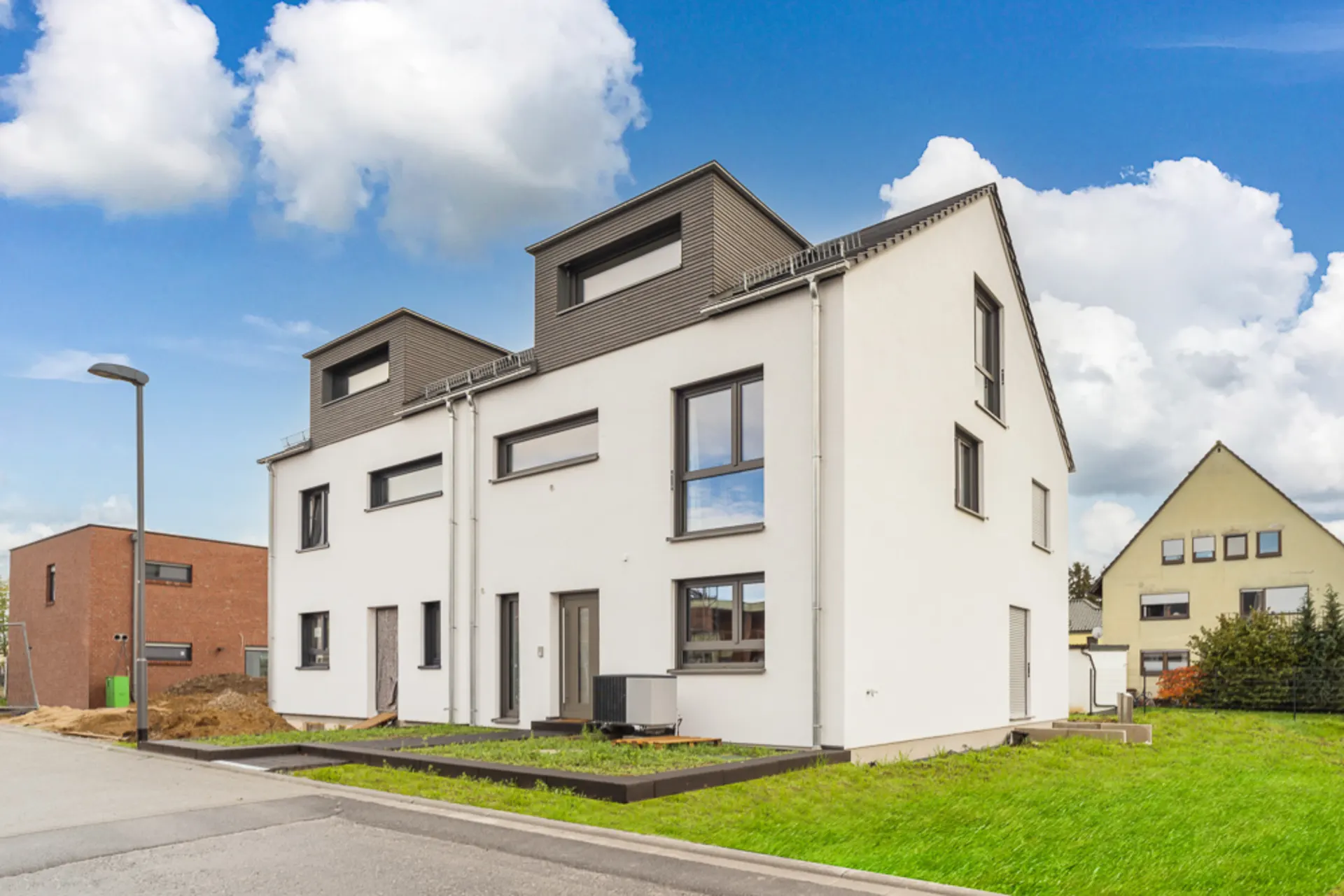 Modernes, weißes Doppelhaus mit grauen Fenstern und zwei Dachgauben, auf grüner Wiese in Wohngebiet.