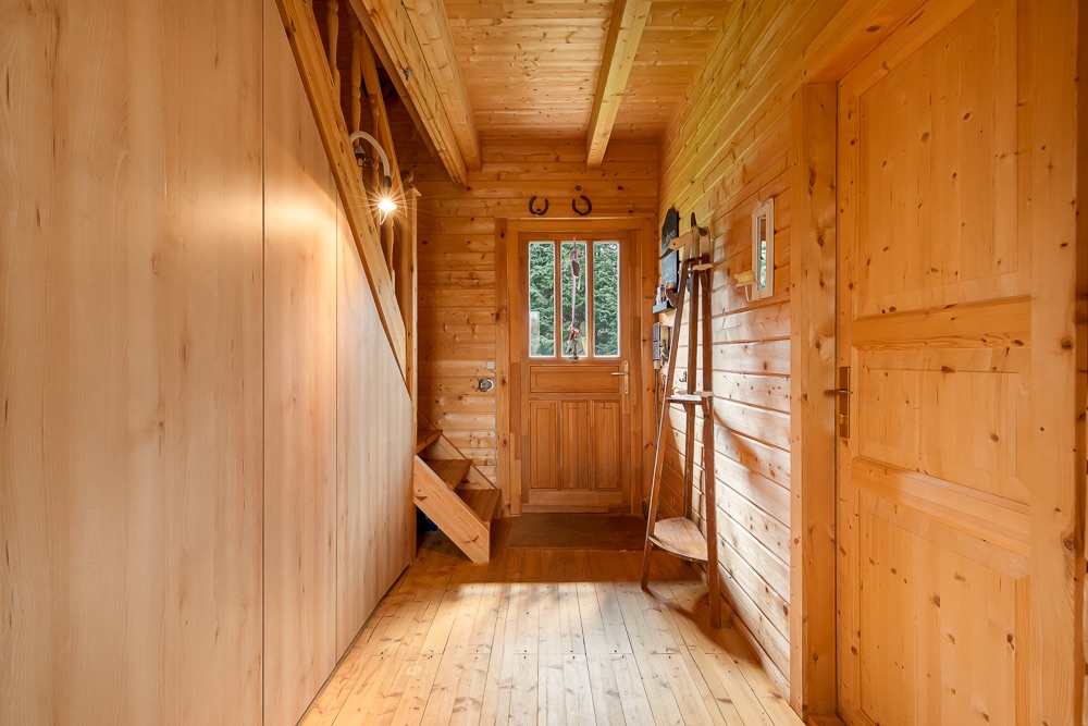 Flur mit Holzwänden, Holztür und Fenster, links eine Treppe, rechts eine geschlossene Tür.