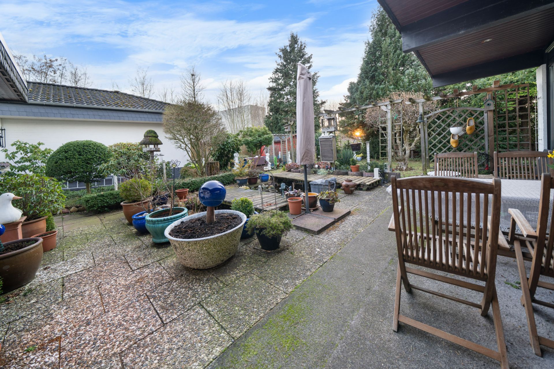 Gepflegter Garten mit vielen Blumentöpfen, Holzmöbeln und Sonnenschirm auf einer Terrasse.
