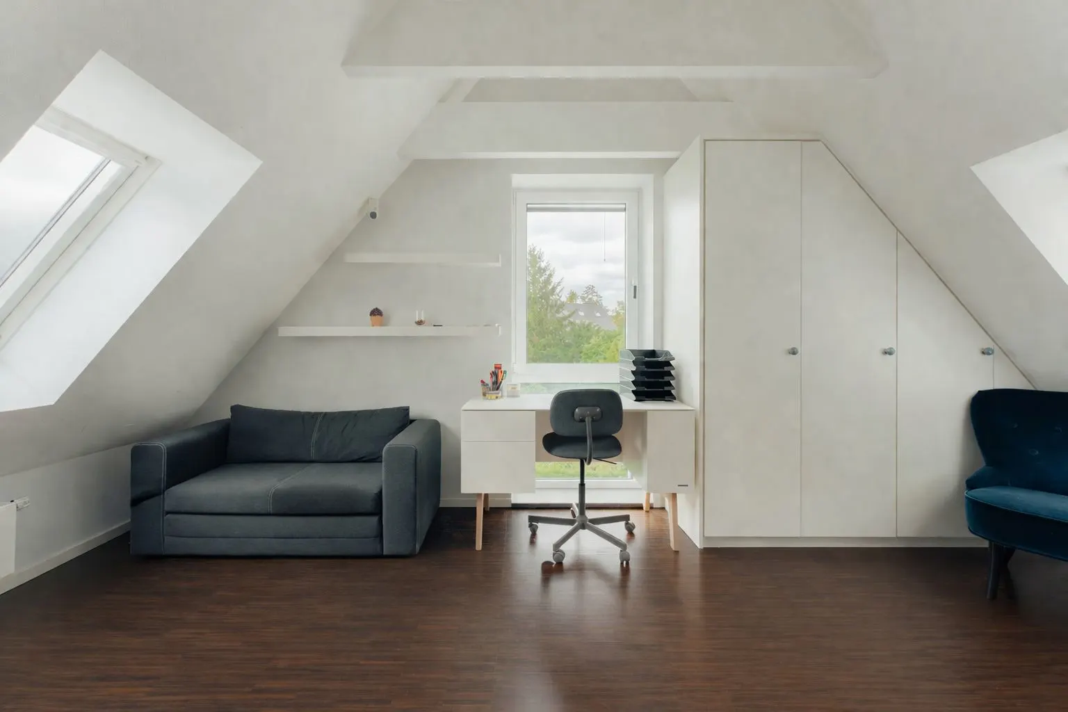 Helles Dachzimmer mit Sofa, Schreibtisch, Stuhl, Regalen und weißen Einbauschränken.