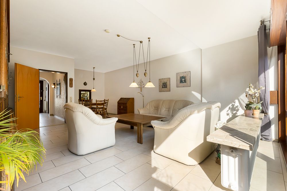 Helles Wohnzimmer mit weißen Sofas, Holztisch und großen Fenstern, dekoriert mit Pflanzen und Bildern.