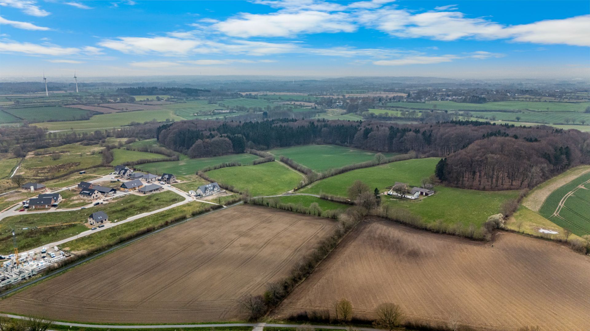 Luftaufnahme von Feldern, Waldflächen und verstreuten Häusern unter blauem Himmel.
