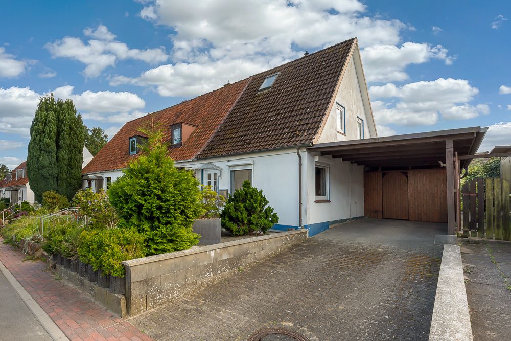 Einfamilienhaus mit rotem Dach, Einfahrt und überdachtem Carport, umgeben von Pflanzen.
