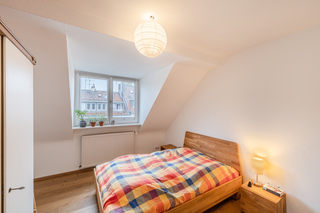 Helles Schlafzimmer mit Holzbett, bunter Bettdecke und Pflanzen auf dem Fensterbrett.