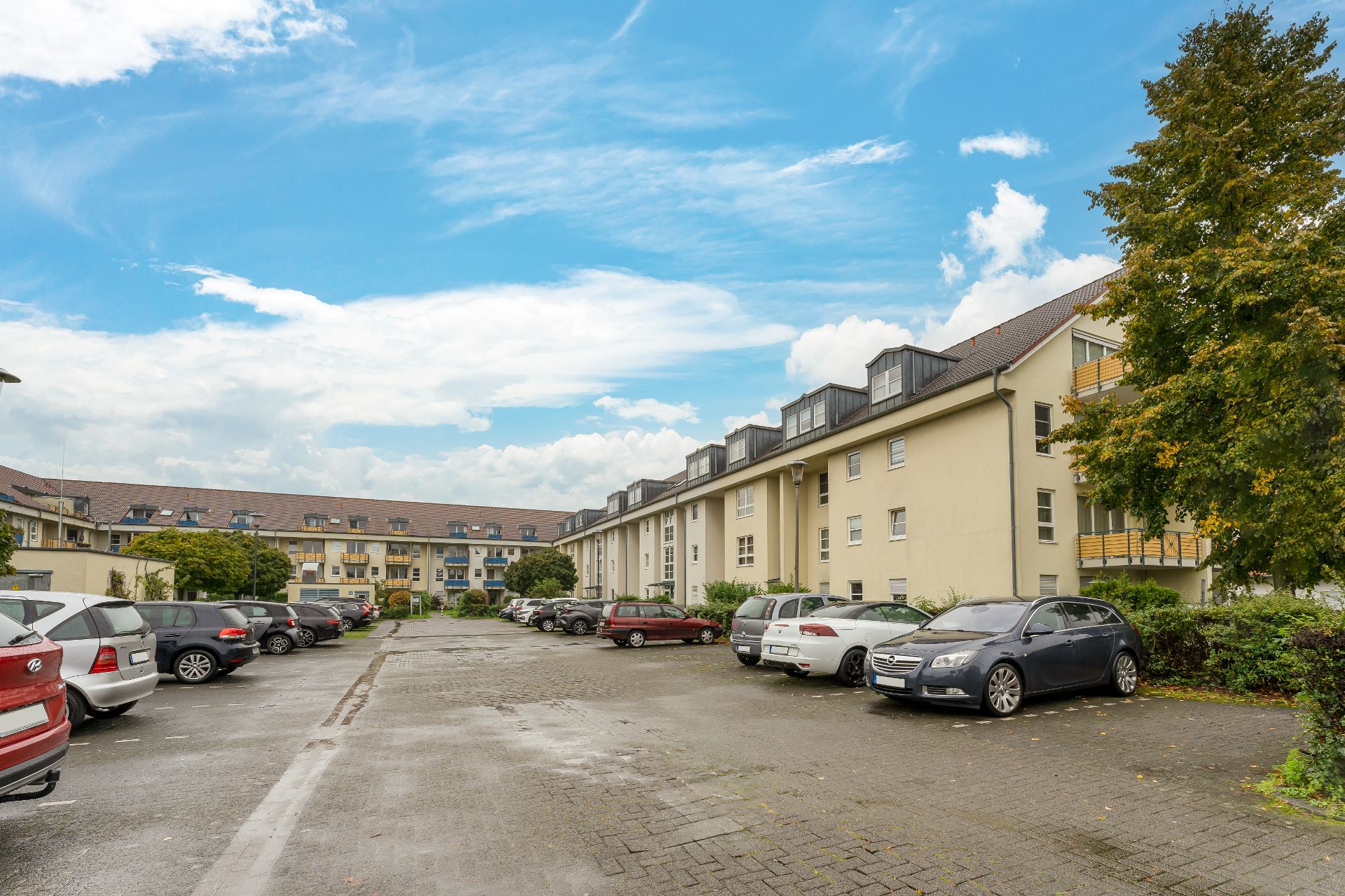 Mehrfamilienhaus mit geparkten Autos auf einem Parkplatz, Bäume und blauer Himmel im Hintergrund.