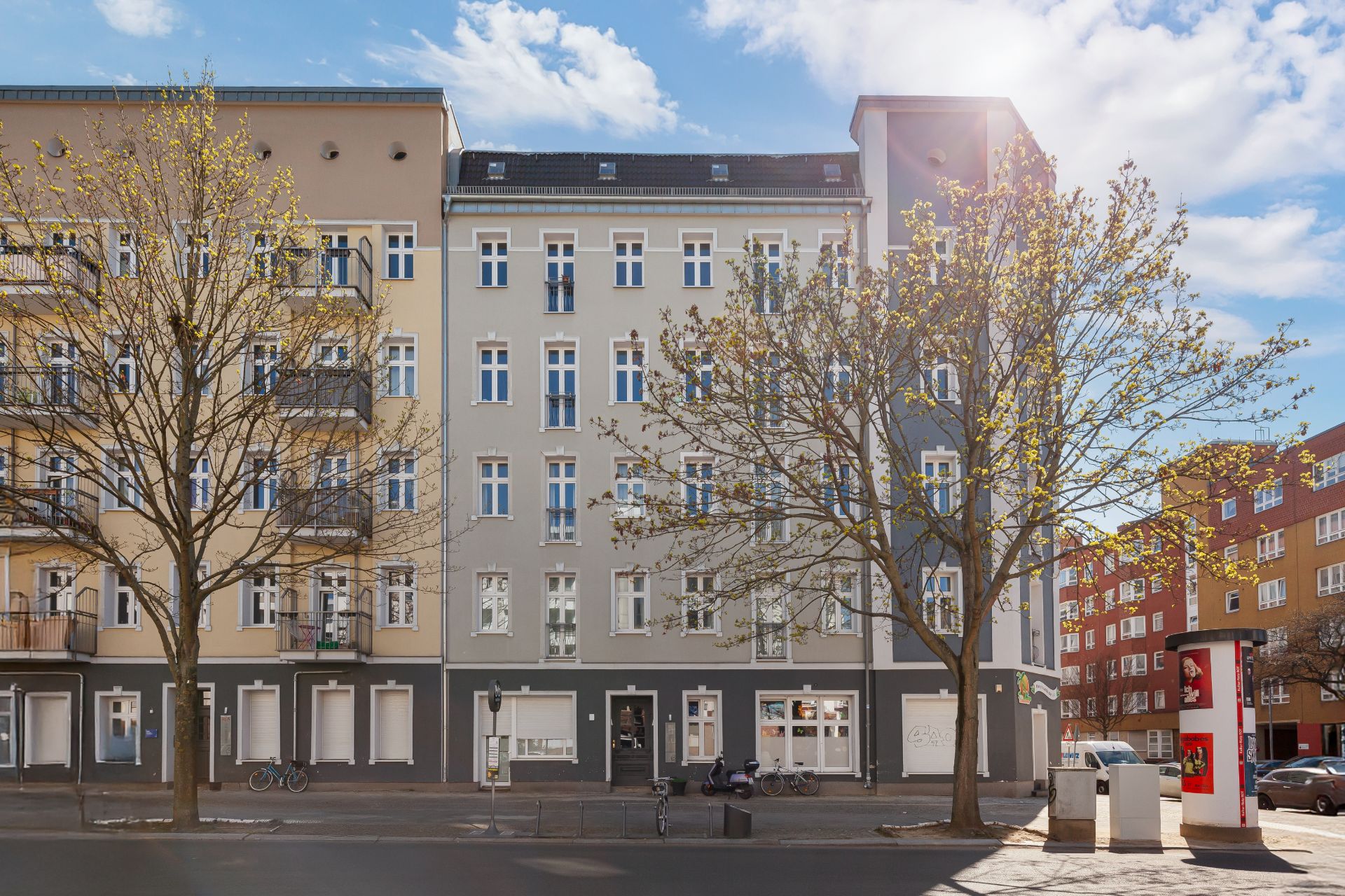 Mehrstöckiges Wohnhaus mit grauer Fassade, großen Fenstern und zwei Bäumen davor an einer Straße.