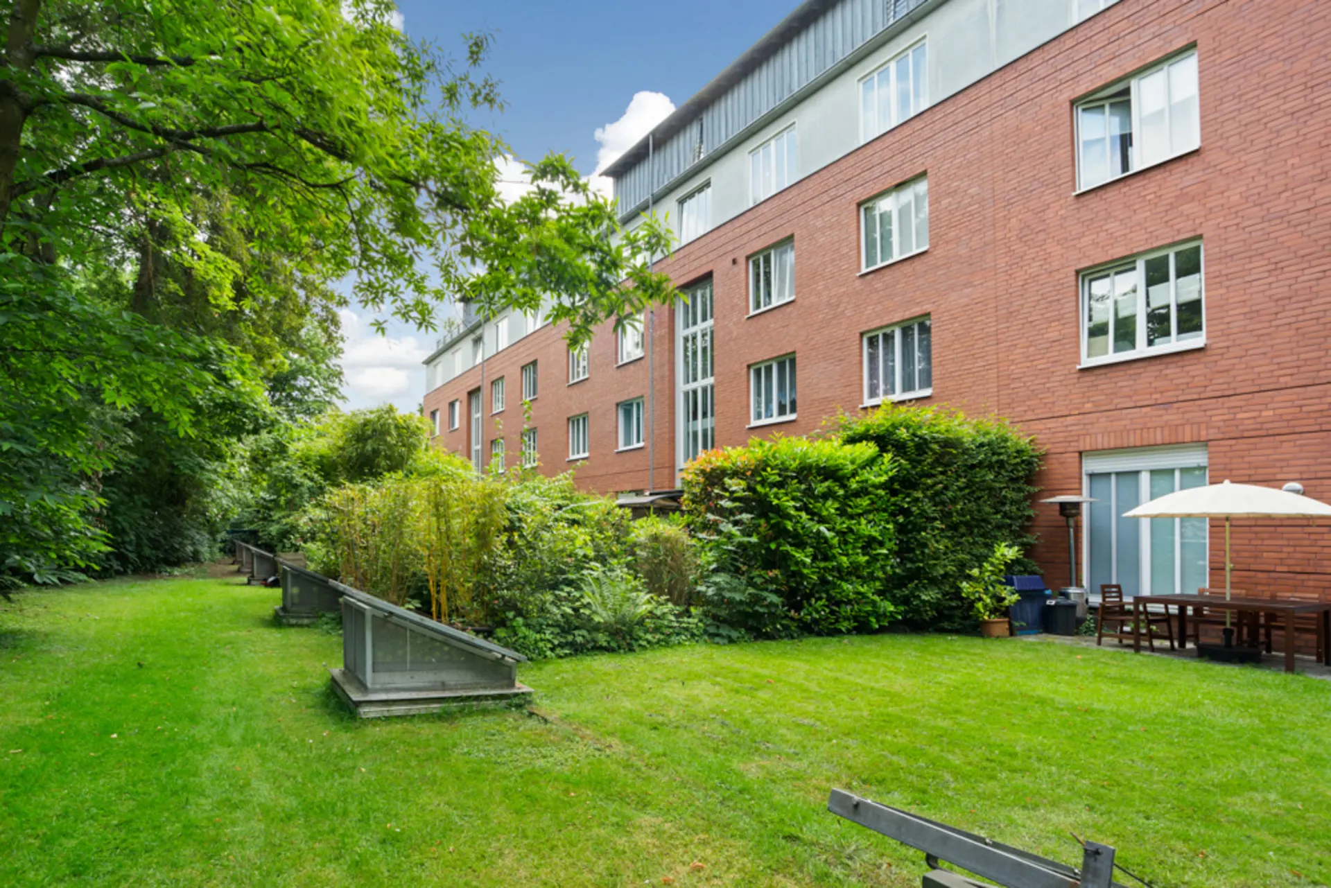 Roter Backsteinwohnblock mit Garten, Rasen, Sträuchern und Sitzbereich mit Sonnenschirm.