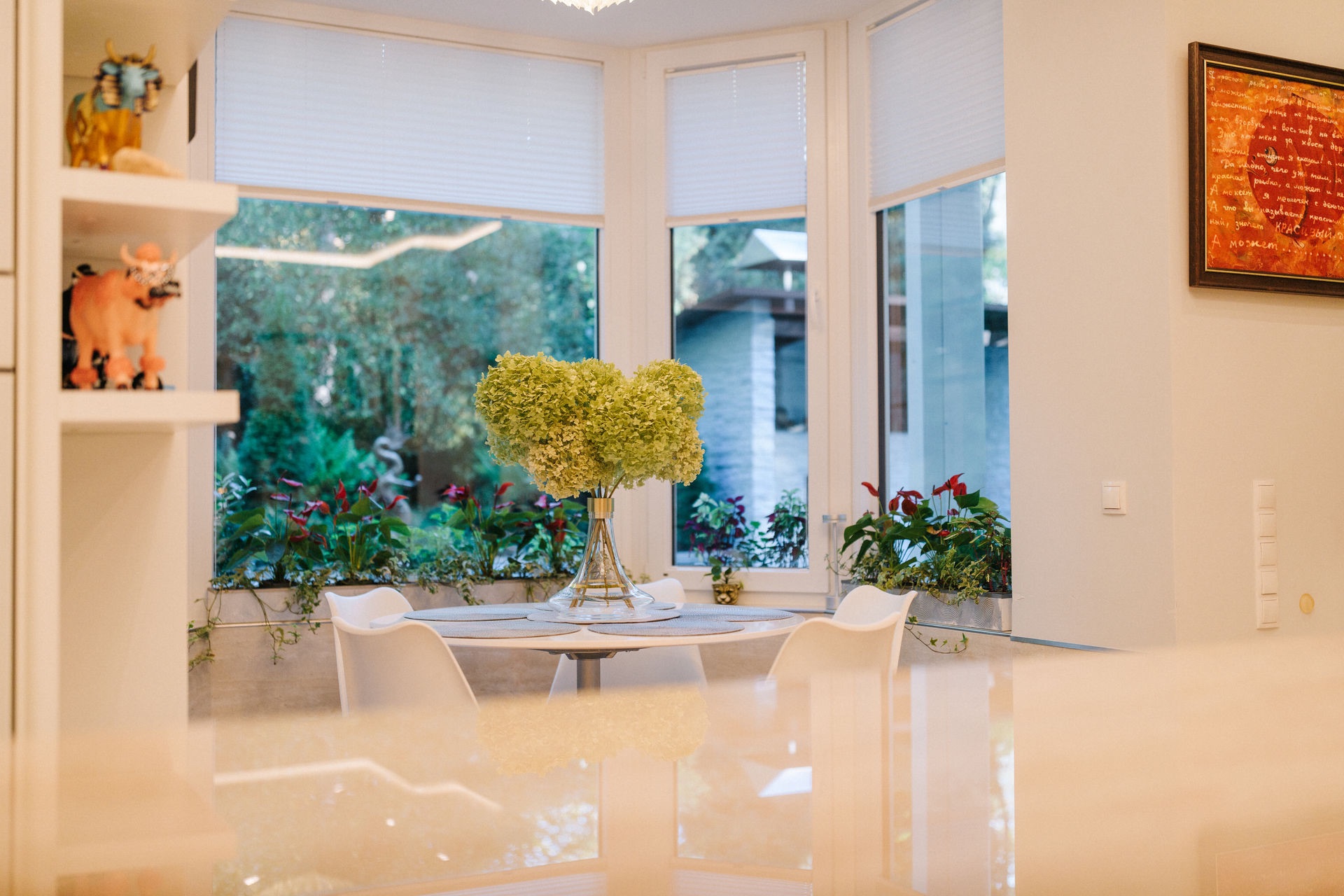 Essbereich mit rundem Tisch, vier weißen Stühlen und großem Blumenstrauß vor großen Fenstern.